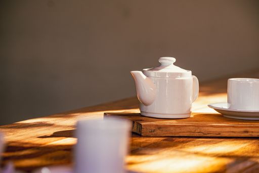 A closeup view of a white cup and a teapot placed on a wooden board on a wooden table under sun