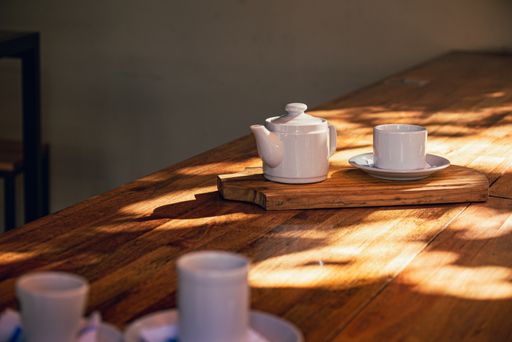 A closeup view of a white cup and a teapot placed on a wooden board on a wooden table under sun
