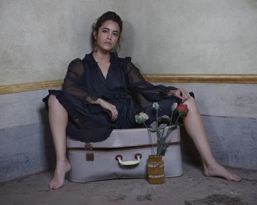A charming and worried woman with tied-up hair is sitting on a ruined suitcase without shoes and wearing a black long dress, watch in camera.