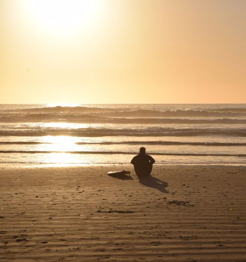 A vertical photo of a silhoette Surfer sitting on the beach next to his Surfboard looking out at the golden sunset and waves
