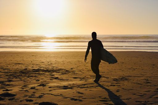 A soft yellow hue photo of a close up silhouette Surfer walking on the beach holding his Surfboard looking at sunset and ocean waves at golden hour