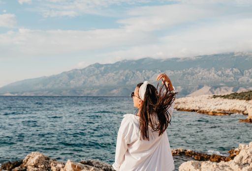 A young Caucasian woman in white long sleeve and sunglasses flipping her hair near the sea