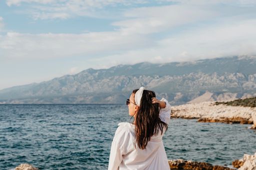 A young Caucasian woman in white long sleeve and sunglasses holding her hair near the sea