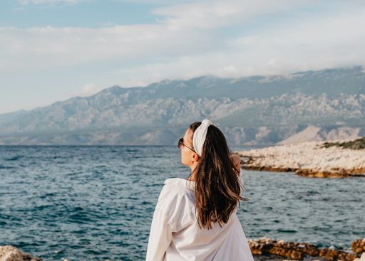 A young Caucasian woman in white long sleeve and sunglasses standing near the sea
