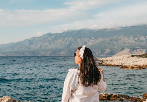A young Caucasian woman in white long sleeve and sunglasses standing near the sea