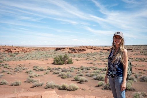 Portrait of a smiling long-haired blonde young girl in a cap in the arid desert in summer, she is carrying her backpack and photo camera