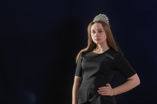 A young girl in the image of a princess with a diadem on her head stands against a dark background and looking into the camera, hand on the waist