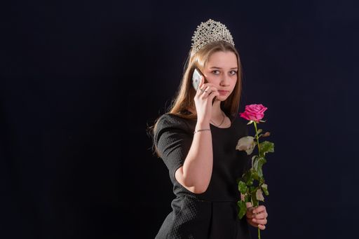 Young girl in the image of a princess with a diadem on her head is standing against a dark background and talking on the phone. Looks into the camer