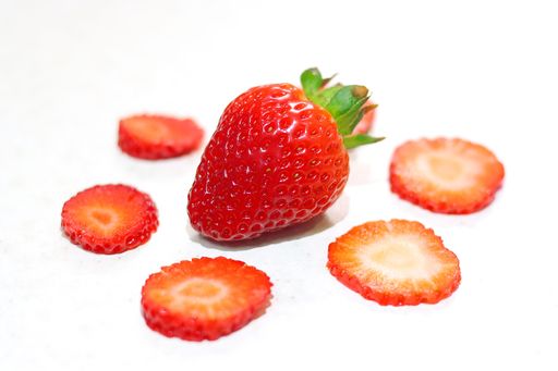 A single, large, fresh, red strawberry surrounded by sliced strawberries on a white surface
