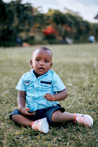 A young Ugandan baby boy in a blue shirt sitting on the grass.