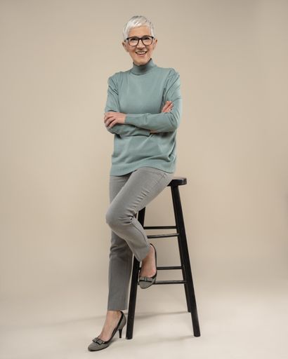 A portrait of an adult woman with short white hair and glasses wearing a turtleneck and posing in a studio