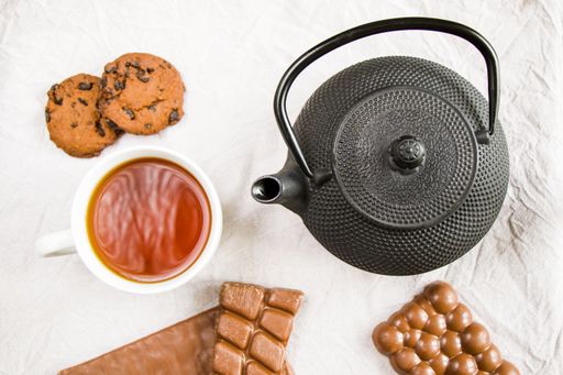 A cup of tea, an iron teapot, cookies, and chocolate bars on a white cloth background