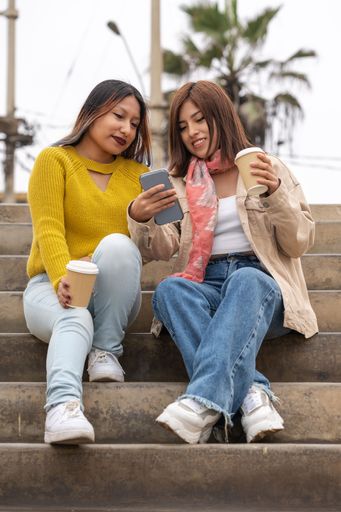 Vertical photo of two friends drinking a take away coffee while using a mobile sitting on the street