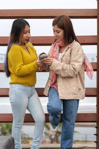 Vertical photo of two latin female friends using a mobile while standing in the street
