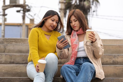 Two latin friends drinking a take away coffee while using a mobile sitting on the street