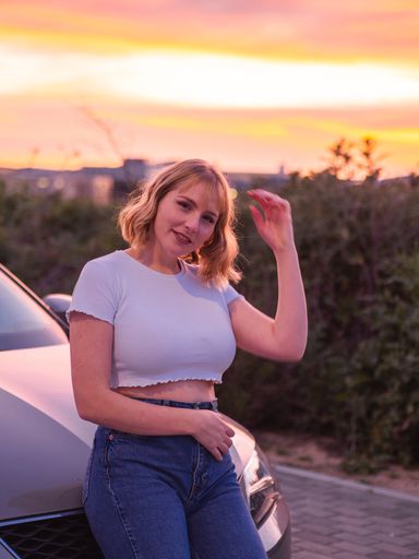 A shallow focus shot of a blonde Caucasian female with a white shirt standing in front of the car at sunset