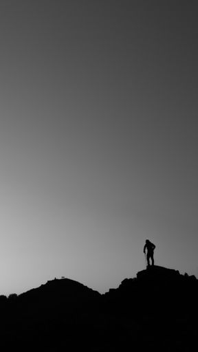 A silhouette of a man standing on top of a rocky hill in grayscale - great for wallpaper or background