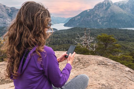 A beautiful brunette Latina woman typing on her phone while sitting at the top of the rocky mountain