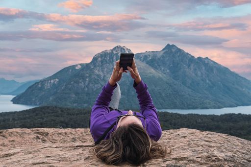 A beautiful brunette Latina woman typing on her phone while lying on a rocky mountain