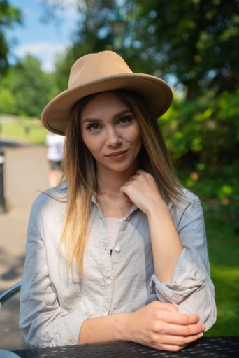 A young beautiful blonde woman in a wide-brimmed hat and pale gray shirt looking at the camera.