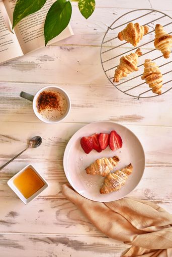 A breakfast set with four freshly-baked croissants, a handful of ripe strawberries, a jar of golden honey, and coffee on a table