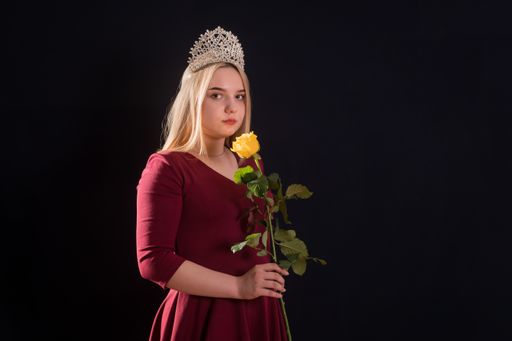 A young blonde girl in a red dress with a diadem on her head holds a yellow rose in her hand