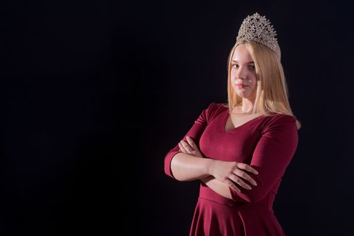 A young blonde girl in a red dress with a diadem on her head folded her arms over her chest and looks into the camera