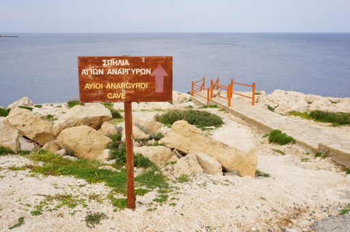 A beautiful shot of an old sign showing the direction to the Anargyroi Cave in front of an ocean
