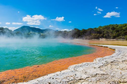 A stunning turquoise pool situated in front of a volcanic crater, with red-hued water and billowing steamy springs