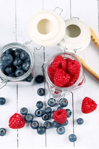 A vertical shot of raspberries and blueberries in jars and a wooden kitchen tool on the table