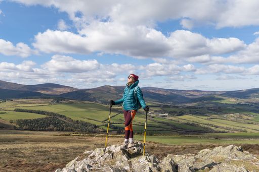 An adult female hiker with trekking poles standing on a rock under a blue cloudy sky