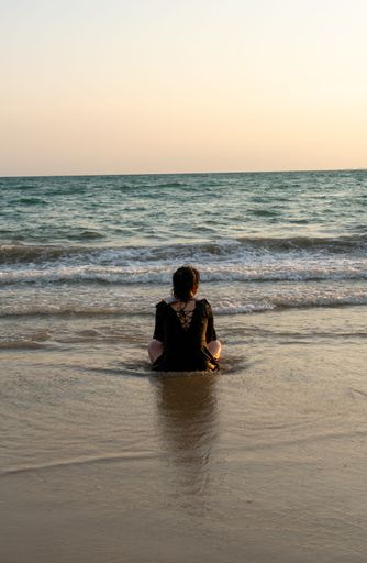 A woman sitting on a sandy shore of a beach, looking as the sun sets in the distance
