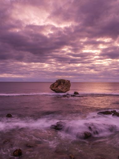 A scenic image of a large rock formation situated on a beach with the ocean at sunset in the background