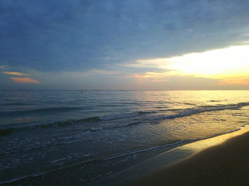 A landscape of a beach surrounded by the sea under a cloudy sky during the sunset
