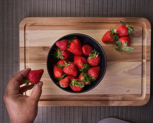 A top view of a person taking a fresh strawberry from a bowl on a wooden cutting board