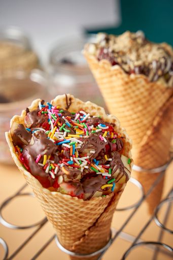 A waffle cone with fruits inside coated in creamy chocolate with colorful sprinkles on top