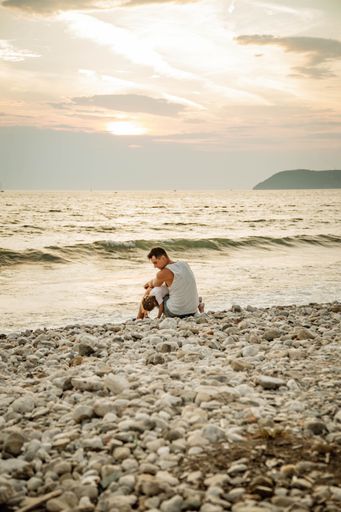 A young adult male with a kid sitting on the sand near the tranquil water on a sunny day