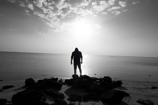 A man standing on a beach in the evening, looking out at the horizon as the sun sets behind a cloudy sky
