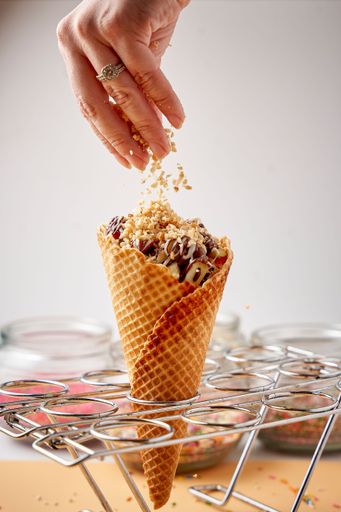 A vibrant image of a woman's hand holding a handful of nutty pieces while pouring them on a freshly-made, colorful ice cream cone