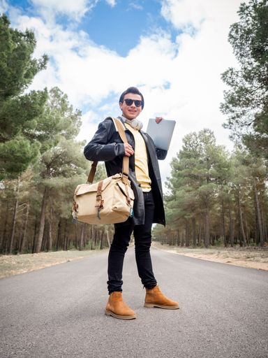 A shallow focus of a young stylish man with a bag and a laptop standing on the road in a forest