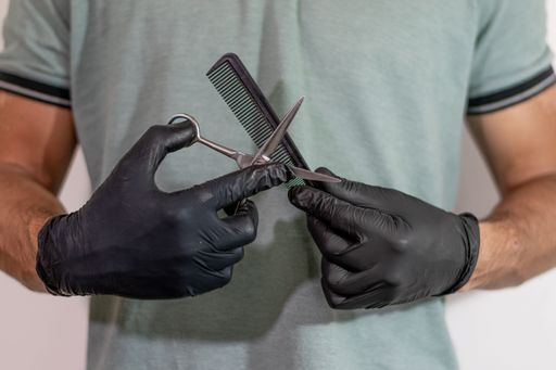 A barber's black-gloved hands holding a pair of scissors to a comb against a white background