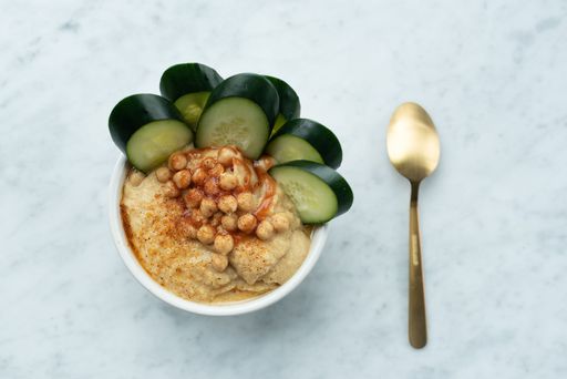 A close-up image of a white ceramic bowl filled with freshly prepared hummus and thin slices of cucumber