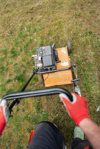 A man wearing protective gloves stands next to a grass mower on a lush green grass field