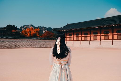 A young female stands in a grand palace courtyard in the fall season, wearing a stunning traditional Korean hanbok dress