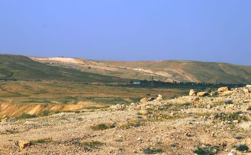 On the foreground, a sandy land of a desert is illuminated by the morning sun. On the background, green and sandy hills meet the bright blue sky.
