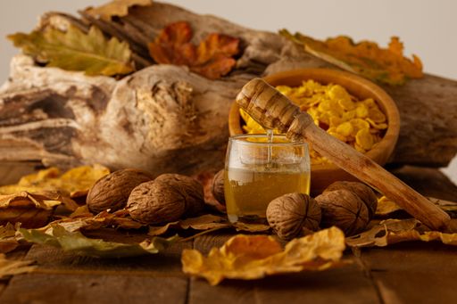 A wooden spoon with dripping honey on a jar, nuts and a bowl of cereal on autumn leaves blurry background