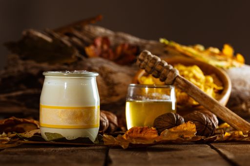 A closeup shot of a wooden spoon on a jar of honey with nuts and a bowl of cereal on a blurry background