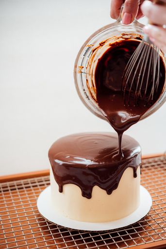 A closeup of a delicious cake with the baker pouring chocolate cream on it