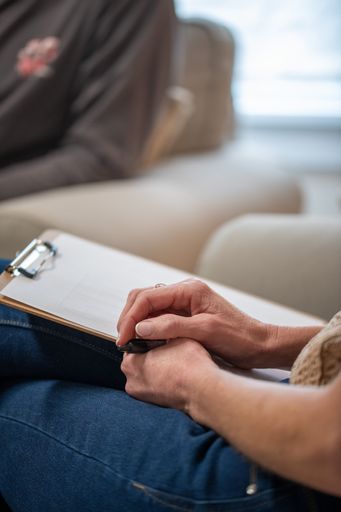 Two people in a professional therapy session, the therapist taking notes as the client talks