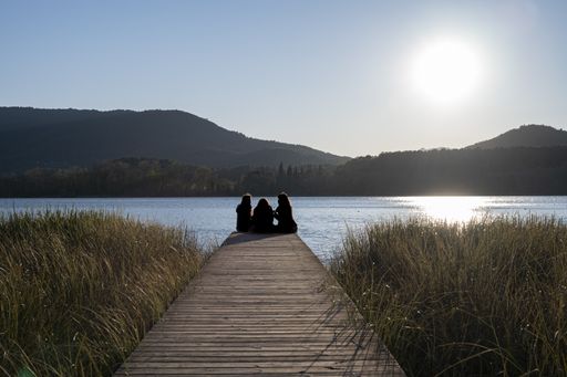 A silhouette of three women sitting on a walkway in the lake with a bright sun in the background